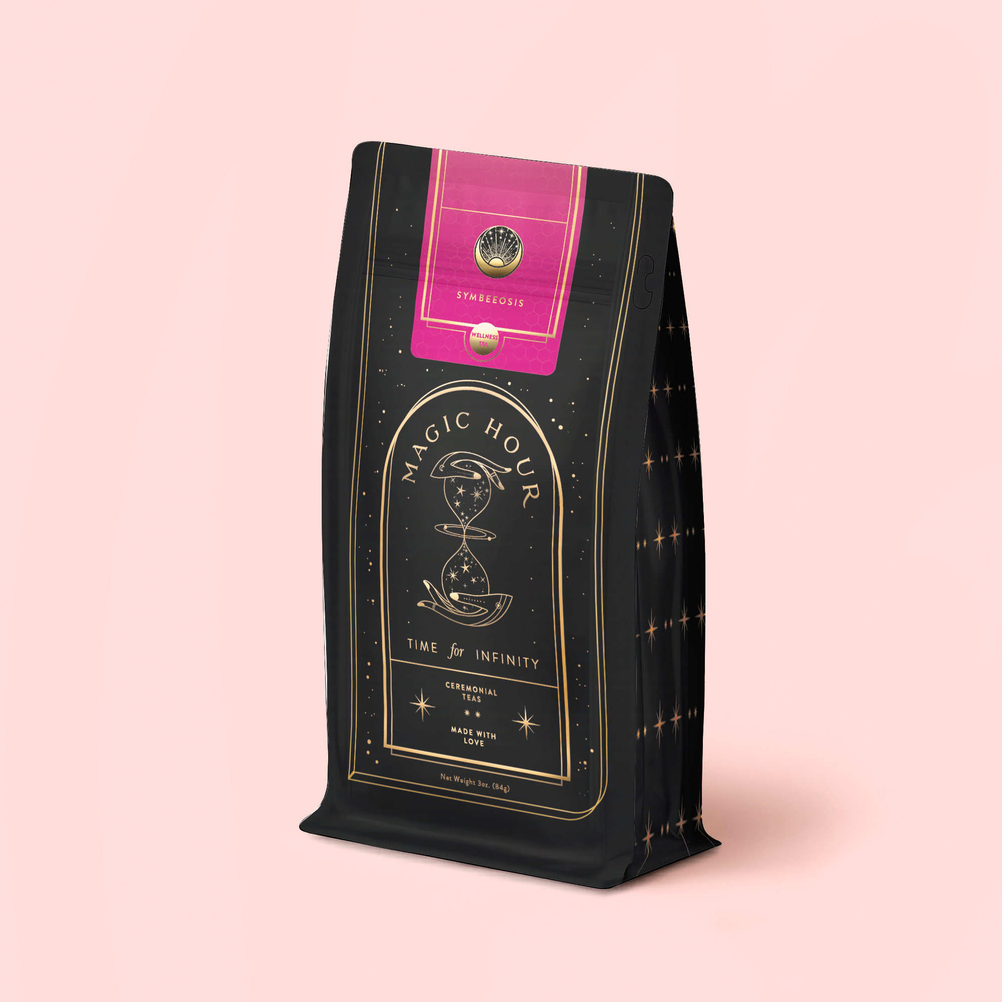 A black and gold "Symbeeosis: Chamomile Elderberry Green Tea Refill Pouch" with celestial designs and a pink-gold label is set against a pale pink background—ideal for enjoying a soothing floral wellness tea blend.