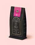 A black and gold "Symbeeosis: Chamomile Elderberry Green Tea Refill Pouch" with celestial designs and a pink-gold label is set against a pale pink background—ideal for enjoying a soothing floral wellness tea blend.