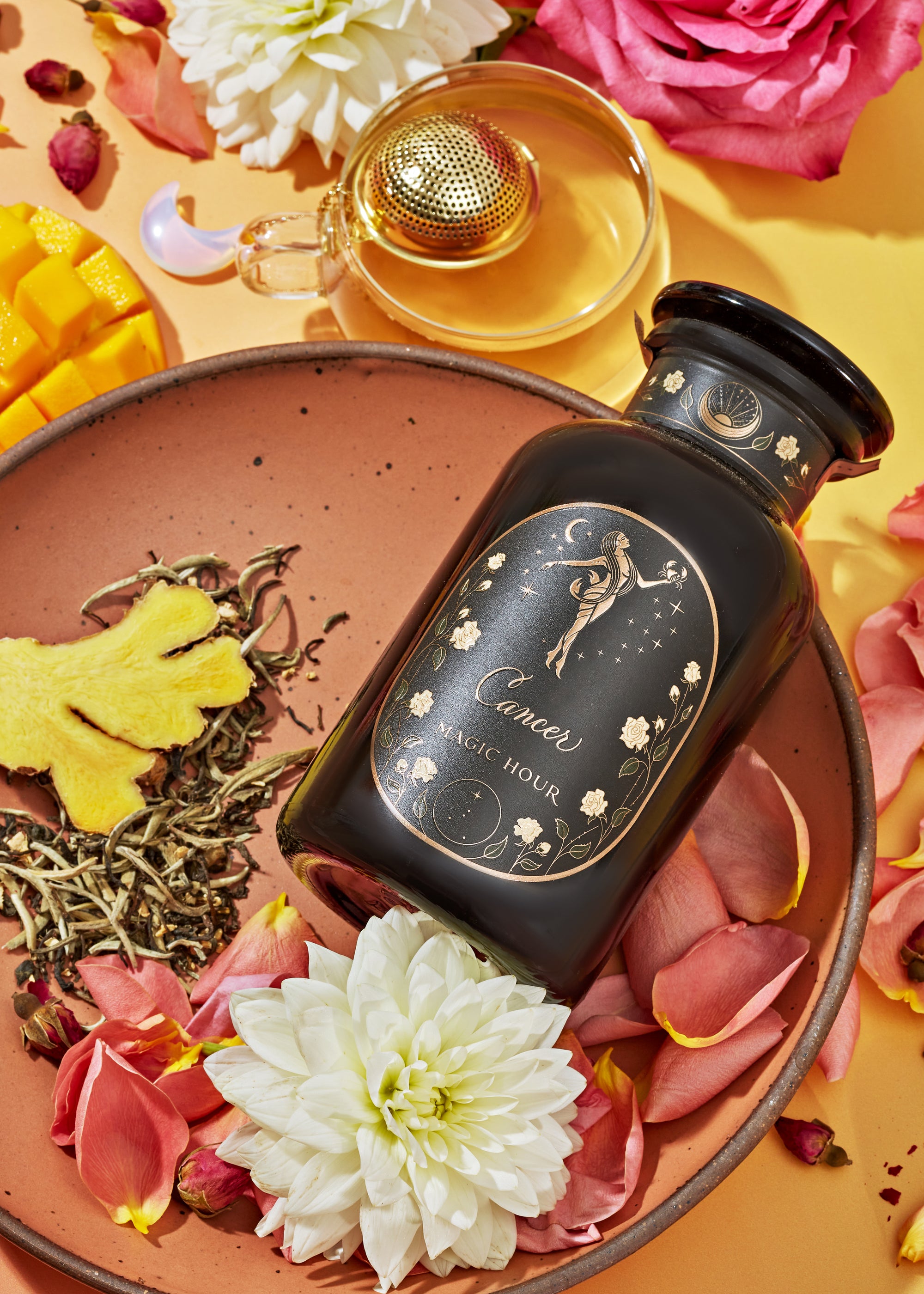 A celestial-themed black jar of tea labeled "Cancer, Magic Hour" is surrounded by white and pink flowers, rose petals, ginger root, a perfume bottle, and pieces of mango on a terracotta plate and a yellow background.