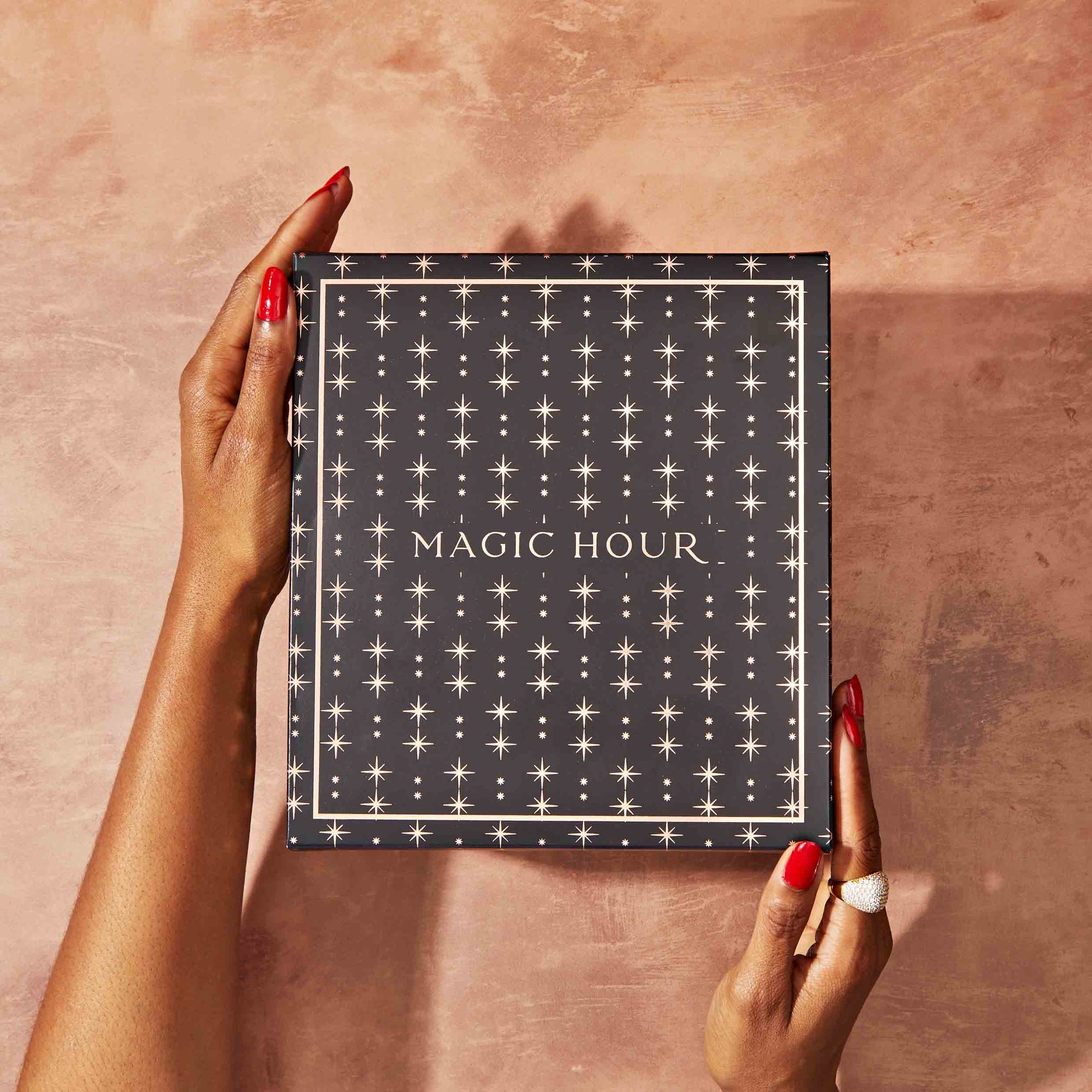 A person with red nail polish and rings holds a dark blue "MAGIC HOUR" box with gold lettering and star patterns, evoking goddess rituals and hinting at the Monthly Magic Tea Journey Subscription, against a textured light brown background.