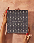 A person with red nail polish and rings holds a dark blue "MAGIC HOUR" box with gold lettering and star patterns, evoking goddess rituals and hinting at the Monthly Magic Tea Journey Subscription, against a textured light brown background.