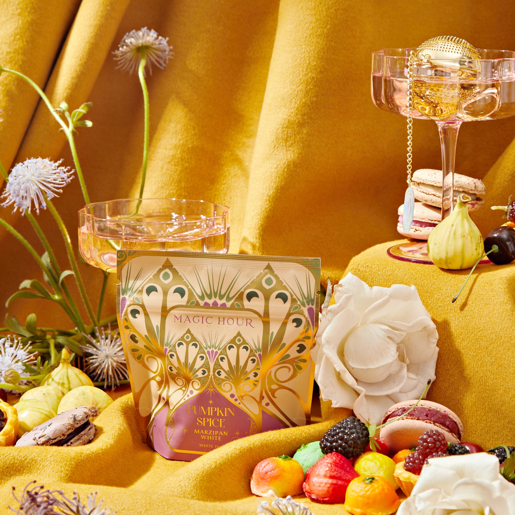 A whimsical still life scene with a box of The Best of Fall Box as the focal point. Around it are ornate glass goblets, assorted macarons, fruit, and white flowers, all set against a backdrop of rich yellow fabric with delicate floral accents from The Best of Fall Box.