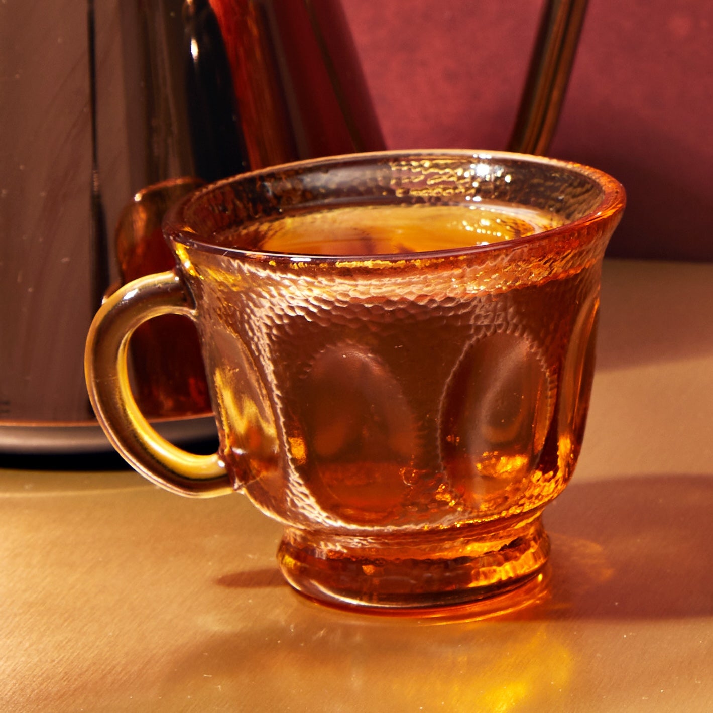 A textured amber glass cup filled with Queen of the South Herbal Tea sits on a gold surface, showcasing its rich adaptogenic blend, with a blurred, similarly colored object in the background.