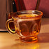 A textured amber glass cup filled with Queen of the South Herbal Tea sits on a gold surface, showcasing its rich adaptogenic blend, with a blurred, similarly colored object in the background.