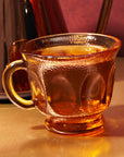 A textured amber glass cup filled with Queen of the South Herbal Tea sits on a gold surface, showcasing its rich adaptogenic blend, with a blurred, similarly colored object in the background.