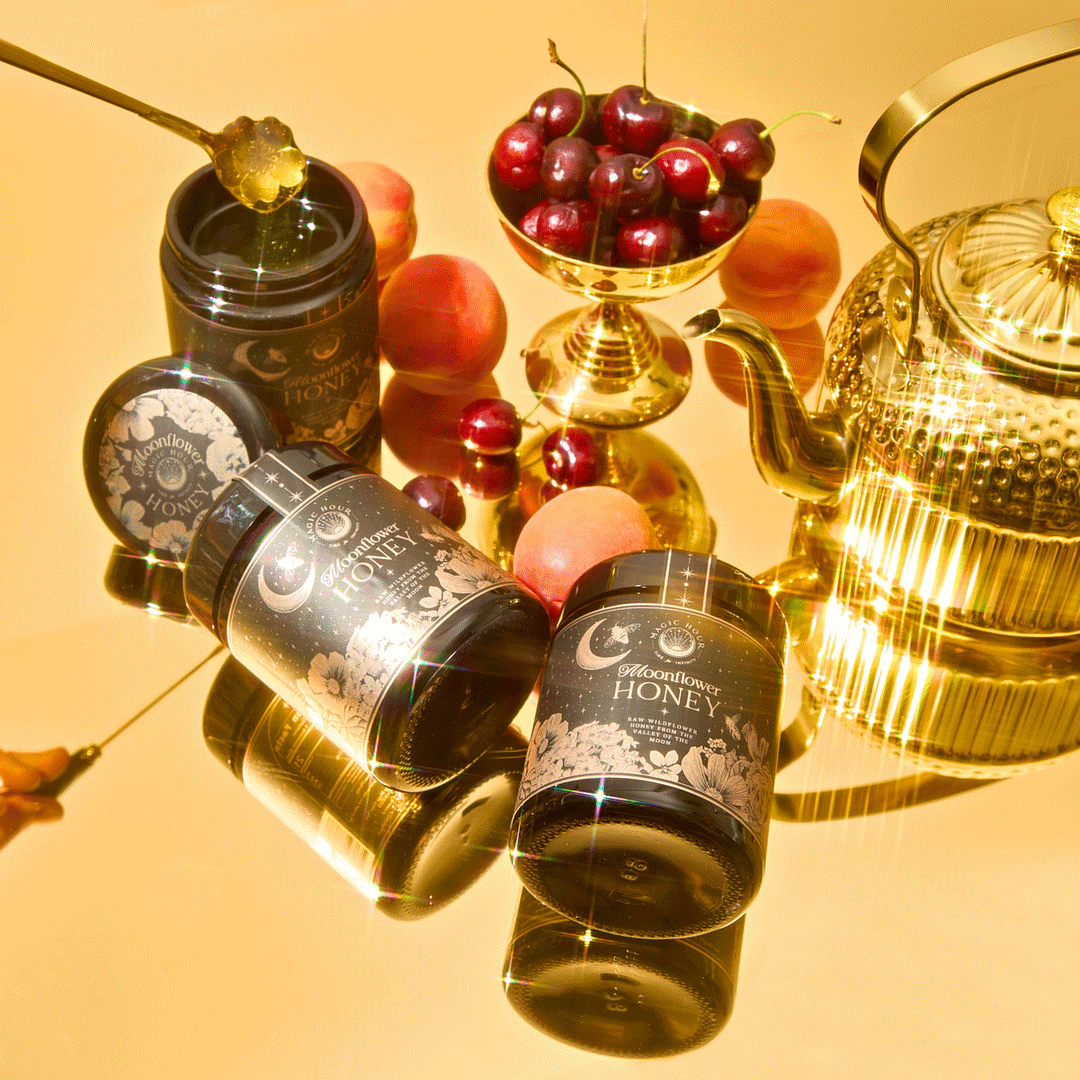 Two jars of Moonflower Honey: 100% Raw, Antioxidant-Rich, Wildflower Honey from Ojai, a bowl of cherries, a gold teapot, and fresh apricots arranged on a reflective surface.
