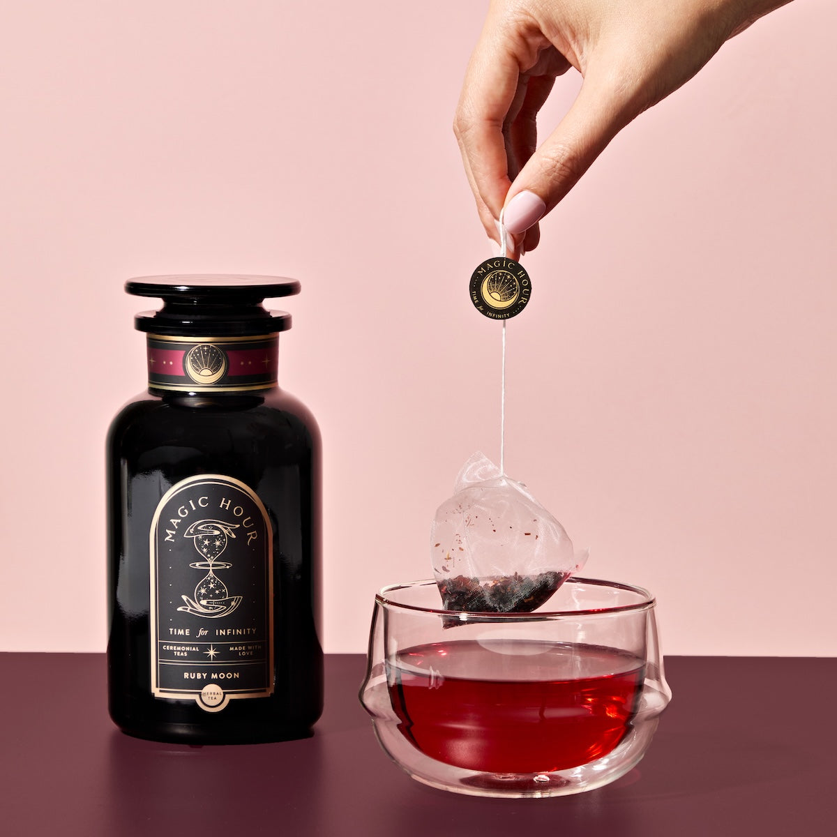 A hand lifts a Ruby Moon™ Herbal Tea Bag from a glass of deep red tea. Beside the cup sits a black jar labeled "Magic Hour," all set against a pink background.