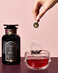 A hand lifts a Ruby Moon™ Herbal Tea Bag from a glass of deep red tea. Beside the cup sits a black jar labeled "Magic Hour," all set against a pink background.