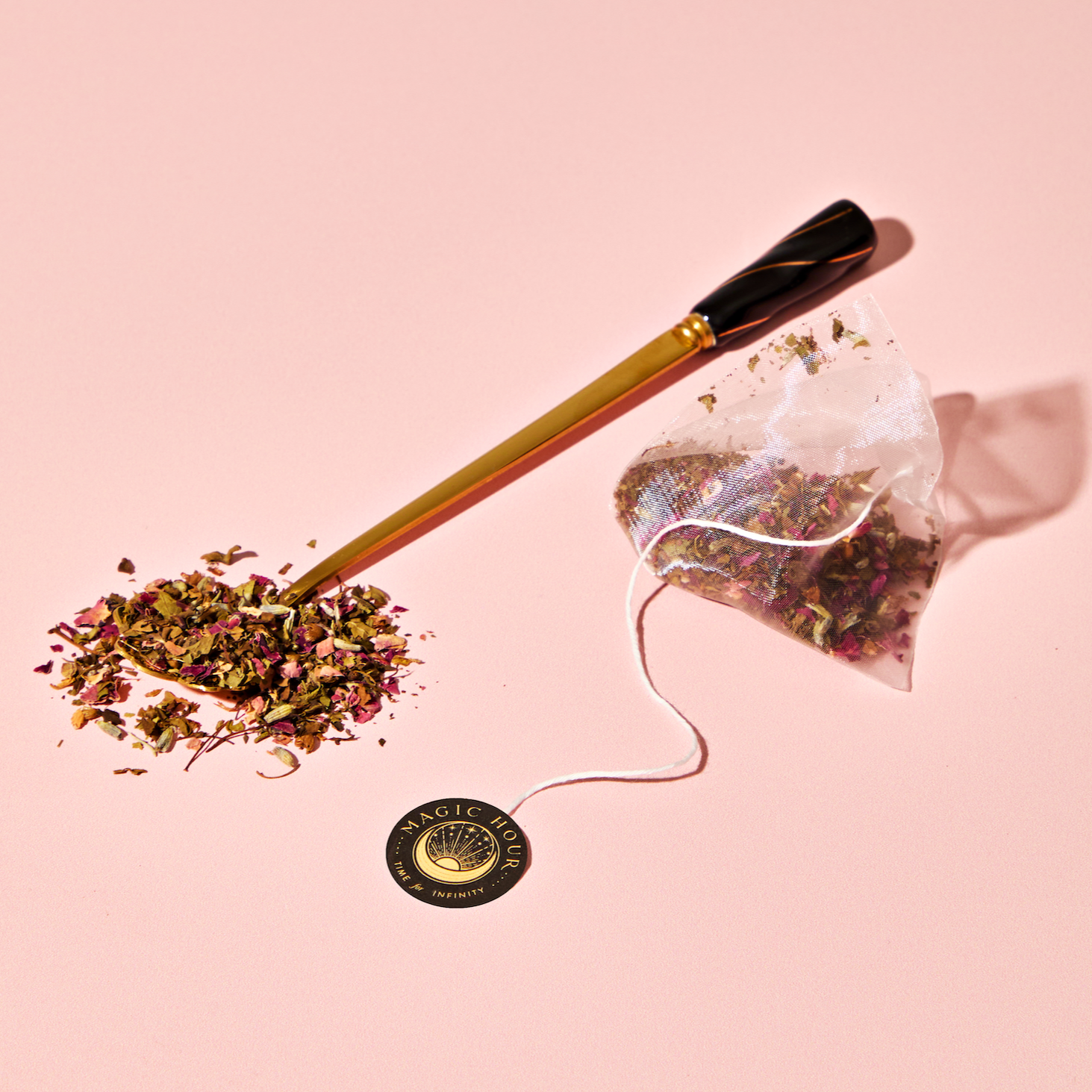 A gold and black spoon holds the Venus – Beauty Sleep Herbal Tea Bags’ botanical herbal blend spilling beside a pyramid tea bag with a round "Magic Hour" label, all set on a pink background.
