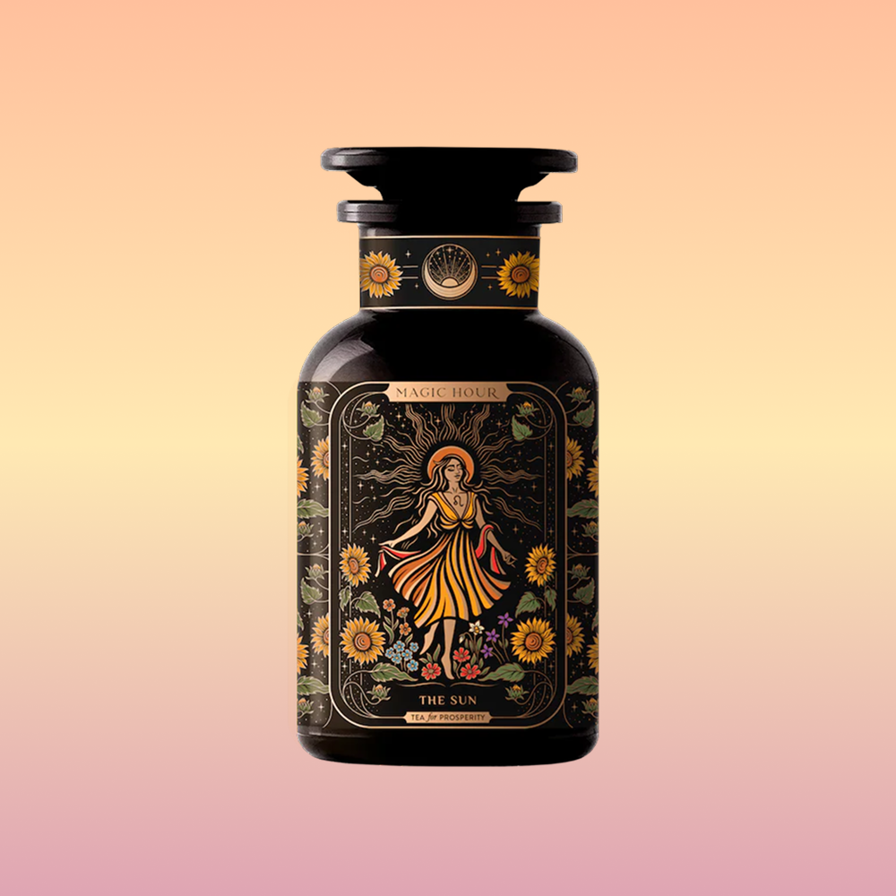 A dark amber bottle from Magic Hour, adorned with ornate label designs, features an illustration of a woman in a yellow dress surrounded by sunflowers and foliage. Titled "The Sun: Energizing Adaptogenic Herbal Tea with Yaupon, Moringa & Turmeric," the bottle has a black cap and displays intricate floral patterns with a mystical theme.