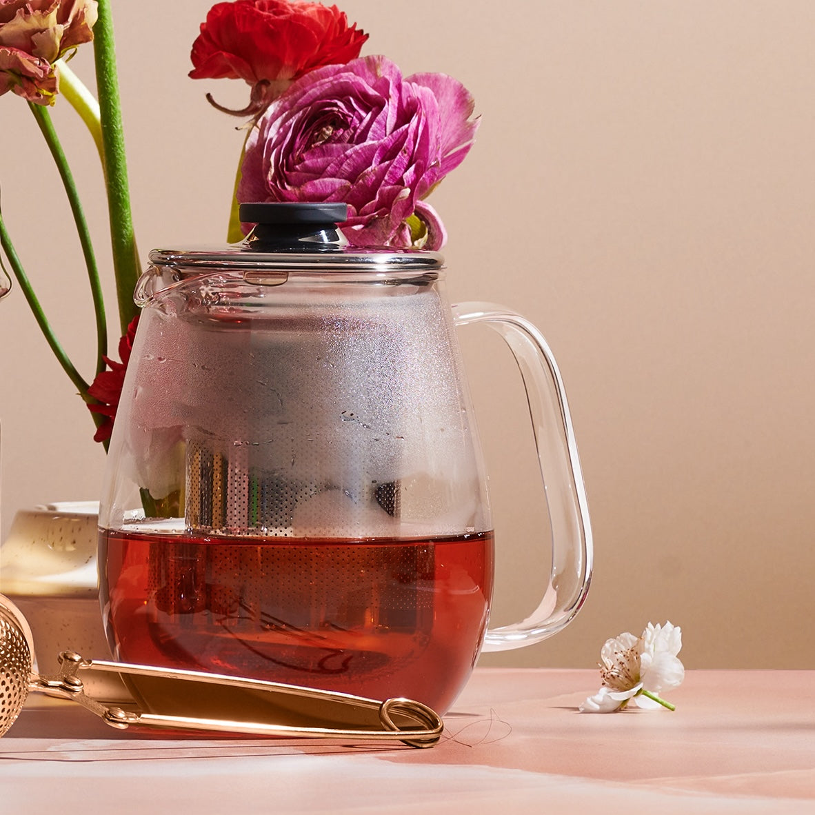 A Magic Hour Kinto Stainless Steel Teapot filled with organic tea sits on a table. The teapot has a stainless steel infuser inside and a clear lid with a black knob. Beside it is a pair of gold-colored tongs and a small white flower. In the background, there are vibrant pink and red flowers.