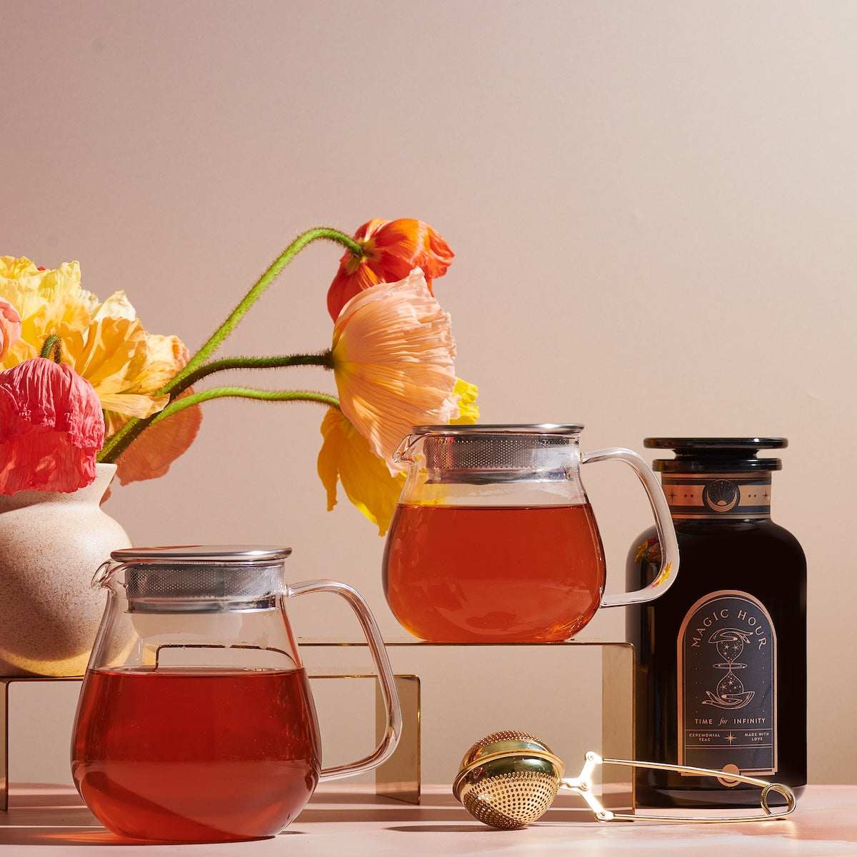 Two Kinto One Touch Teapots filled with organic tea are placed on a table, accompanied by a jar labeled "Magic Hour." A gold tea infuser and string lights lie in the foreground. In the background, there's a ceramic vase with colorful flowers and a soft, warm light illuminating the scene.