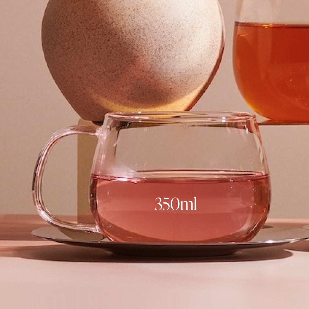 A clear Kinto Glass Tea Cup from Magic Hour, labeled "350ml," sits on a reflective saucer. Behind it, part of another glass filled with organic tea is visible, with a large, round stone in the background. The scene is set on a light pinkish-brown surface.