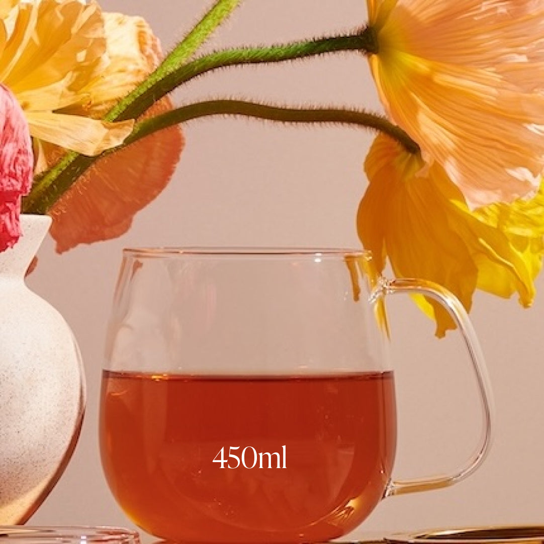 A Kinto Glass Tea Cup filled with a reddish beverage sits beside a white vase holding vibrant yellow and orange poppy flowers. The cup has "450ml" written on it. The soft lighting casts gentle shadows, creating a warm and serene atmosphere, perfect for savoring your Magic Hour tea.