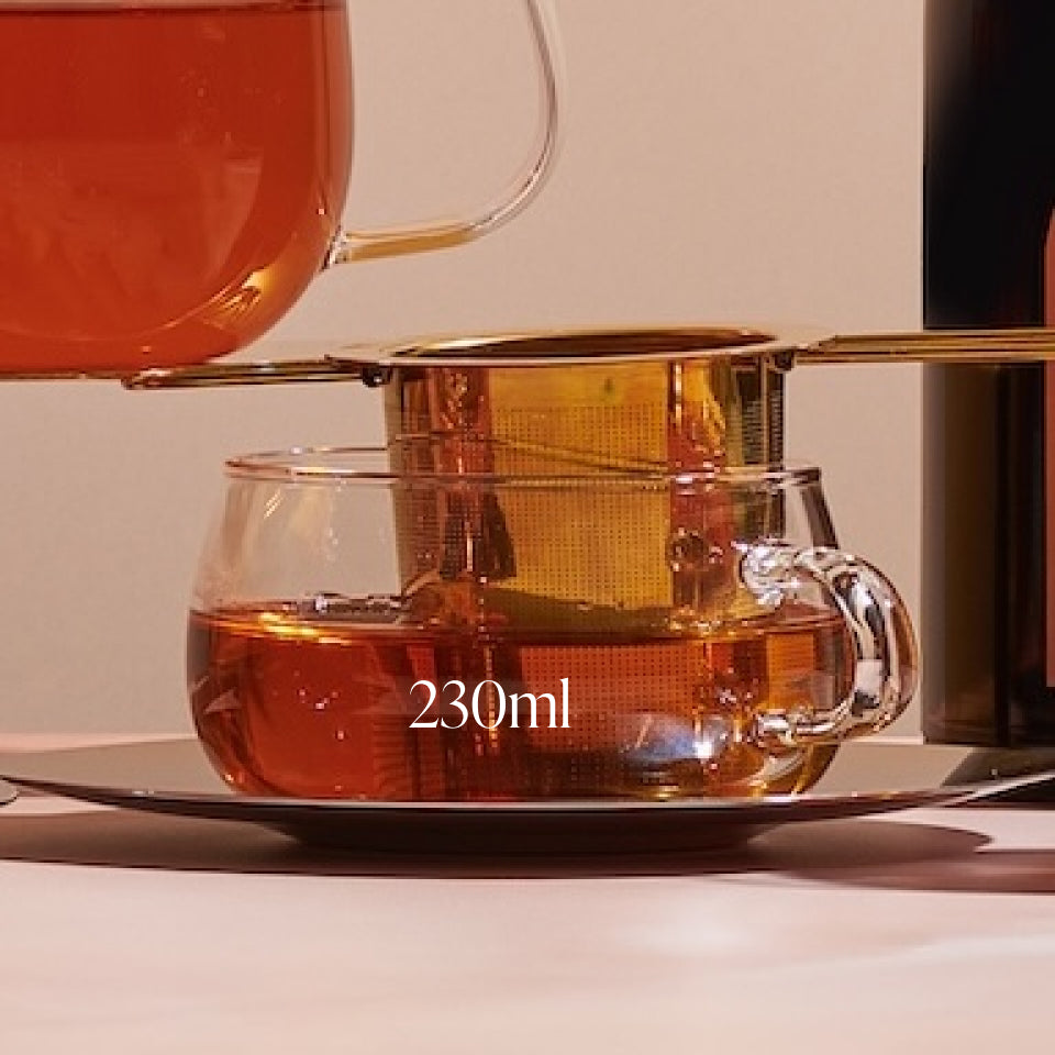 Close-up image of a 230ml Kinto Glass Tea Cup with a metal tea infuser placed on top. Organic tea is being poured from a glass teapot through the infuser into the cup. The setting is warm and inviting, perfect for preparing loose leaf tea during your Magic Hour tea ritual.