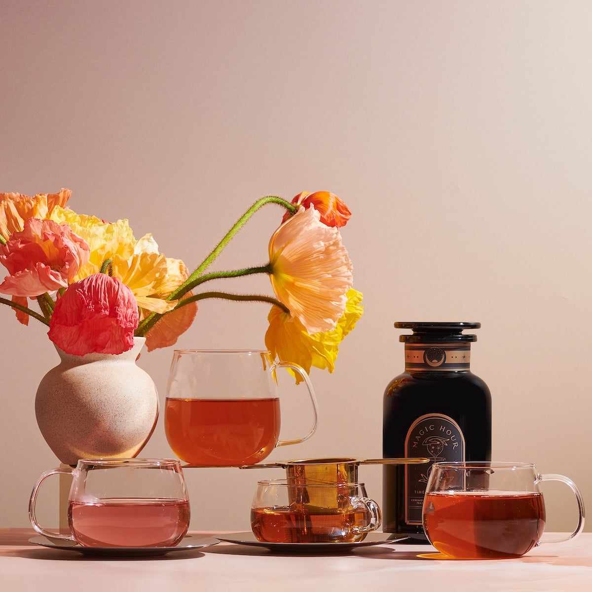 A still life image features a vase with colorful flowers, four Kinto Glass Tea Cups filled with different shades of organic tea, and a bottle of Magic Hour Tea. The background is softly lit with a warm tone, creating a cozy and inviting atmosphere.