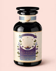 A dark glass jar labeled "Tea for Intuition: 1111: Pineapple & Butterfly Pea Flower Herbal Tea" by Magic Hour, adorned with celestial designs and floral illustrations, rests on a light pink background, evoking enchanting notes of butterfly pea flower.