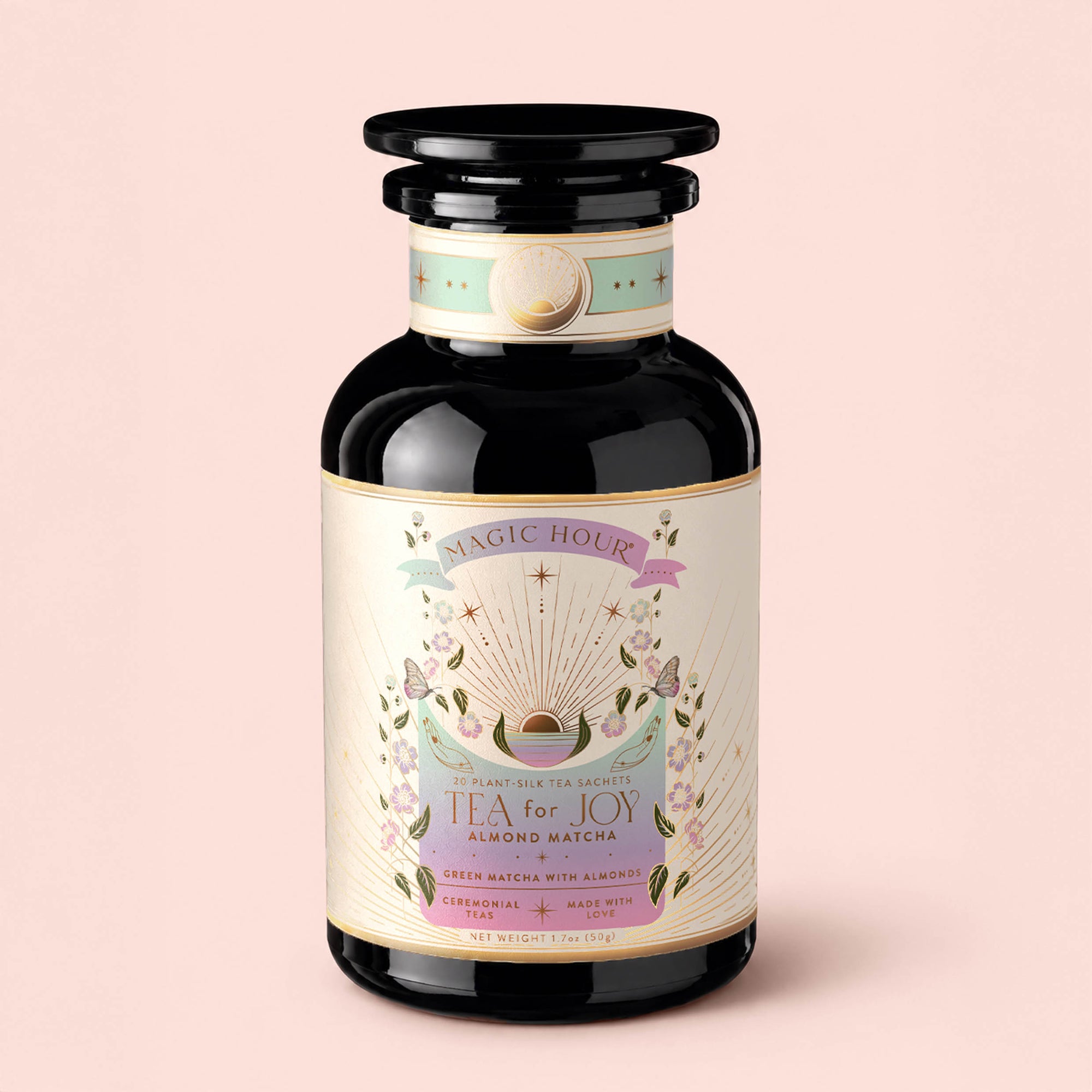 A black glass jar labeled "Tea for Joy: Almond Matcha: Green Matcha Tea with Almonds," featuring pastel floral designs and gold accents, is set against a light pink background. This antioxidant-rich matcha blend delivers a delightful tea experience while naturally supporting metabolism.