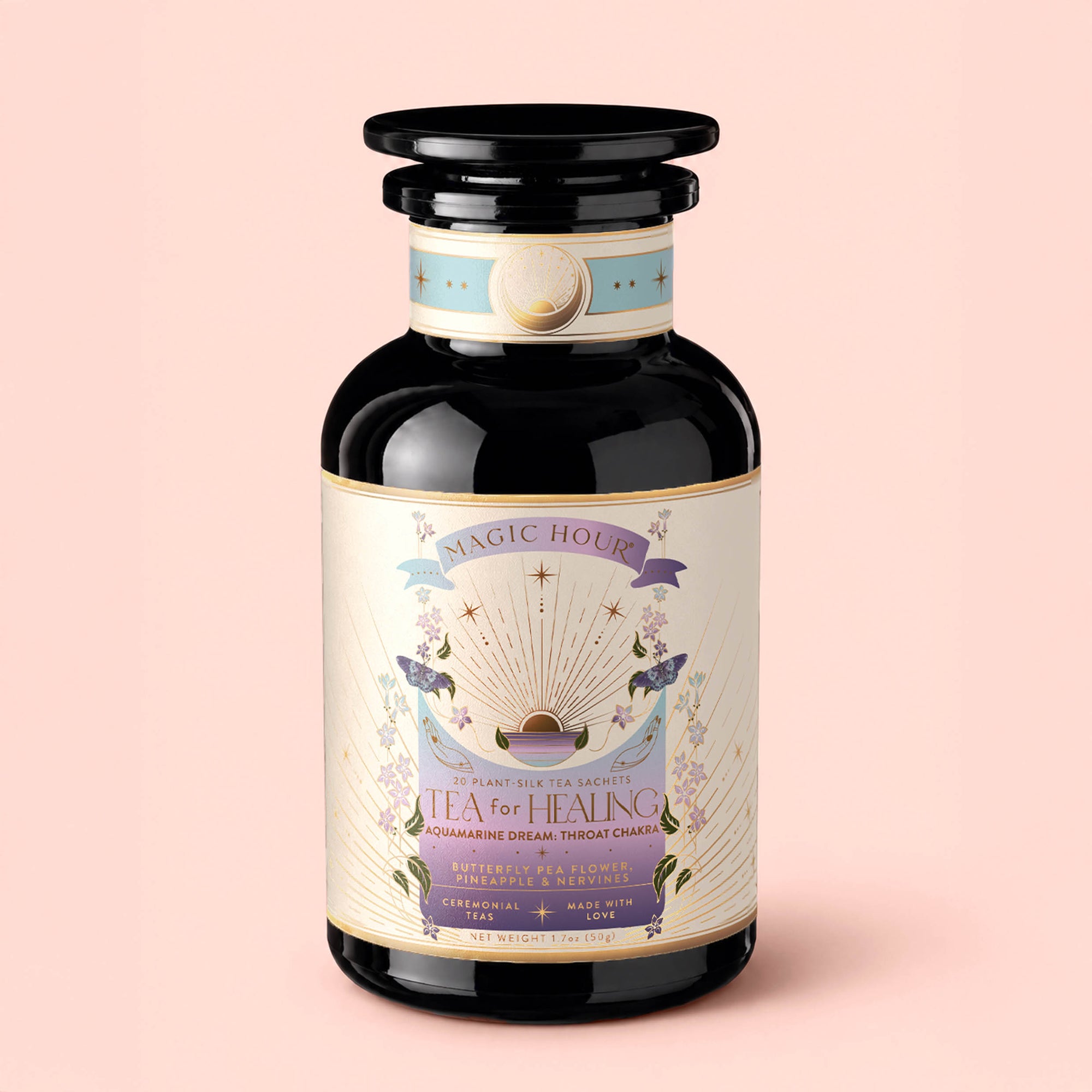 A dark glass jar labeled "Tea for Healing: Aquamarine Dream: Blueberries, Pineapple &amp; Nervines Herbal Tea" rests on a pale pink background. Its label showcases pastel hues, lavender illustrations, and celestial motifs, reflecting its adaptogenic blend designed for nervous system support and restful sleep. The jar features a wide, round lid with gold accents.