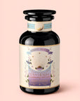 A dark glass jar labeled "Tea for Healing: Aquamarine Dream: Blueberries, Pineapple & Nervines Herbal Tea" rests on a pale pink background. Its label showcases pastel hues, lavender illustrations, and celestial motifs, reflecting its adaptogenic blend designed for nervous system support and restful sleep. The jar features a wide, round lid with gold accents.