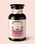 A dark glass jar labeled "Tea for Peace: Child's Pose: Citrus, Lavender & Tulsi Herbal Tea Blend" features a pastel pink and cream design with gold accents, and is described as an adaptogenic tea for the heart chakra, formulated to relax your nervous system—all set against a light pink background.