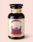 A dark glass jar labeled "Tea for Celebration: Coconut Chai: Rich Spiced Black Tea Blend" features a purple and gold design adorned with stars and radiant rays, evoking crown chakra energy, and is set against a light pink background. The tea inside is a rich spiced black tea blend from Magic Hour.