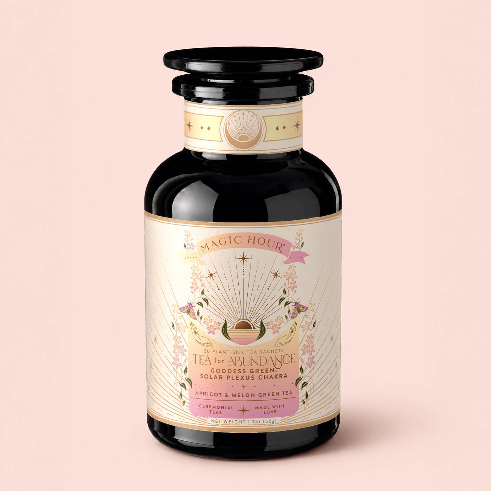 A decorative glass jar labeled "Tea for Abundance: Goddess Green: Apricot &amp; Lychee Green Tea," featuring pastel art, floral accents, and gold details, contains a green tea blend with apricot, lychee, and organic lemongrass by Magic Hour. Known for its refreshing flavor and metabolism-supporting properties, the jar is displayed on a pale pink background.