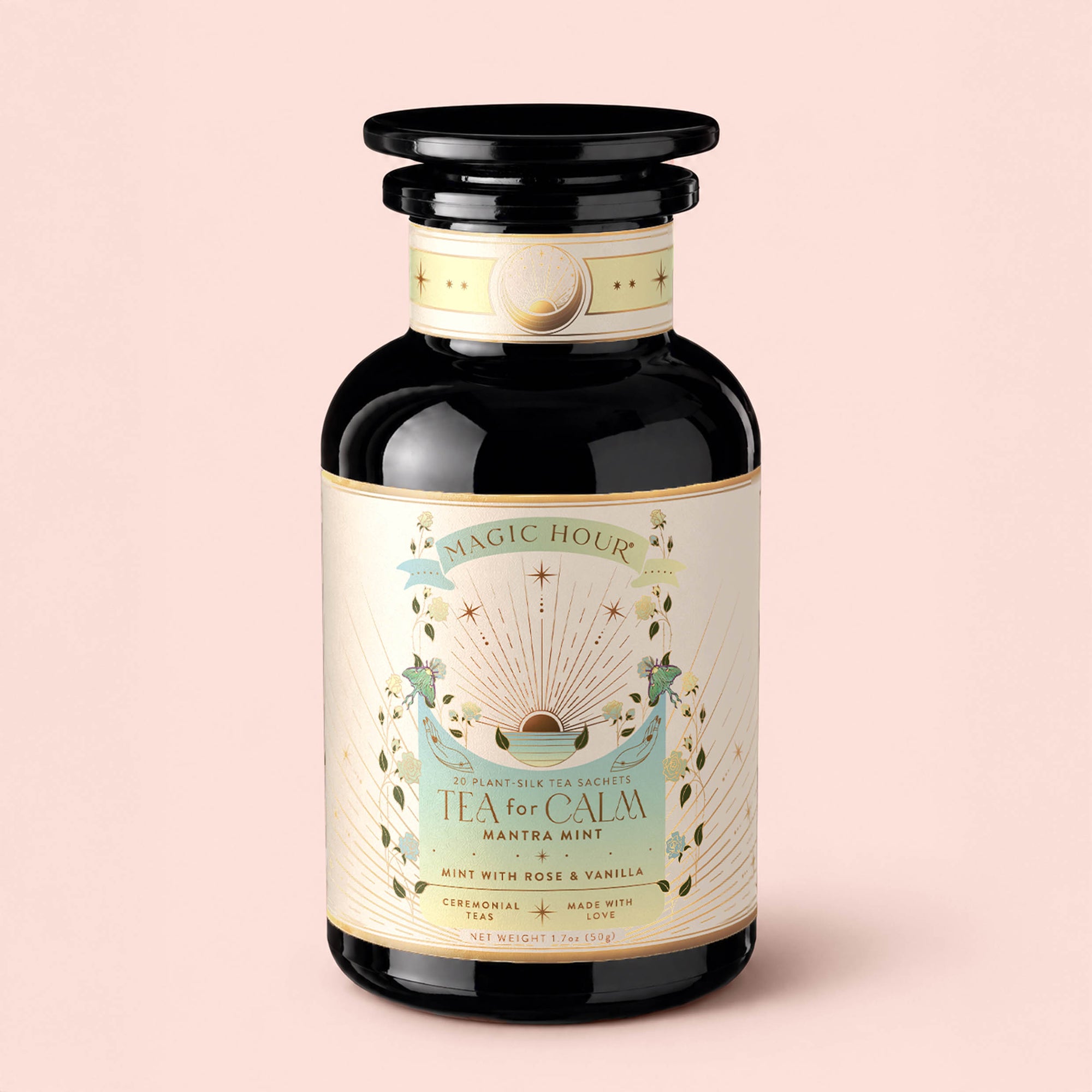 A black jar labeled "Tea for Calm: Mantra Mint: Mint With Rose &amp; Vanilla Herbal Tea," adorned with gold accents, celestial motifs, and botanical illustrations, is set against a light pink background. This organic herbal tea blend features soothing notes of mint, rose, and vanilla, making the Mantra Mint Herbal Tea ideal for moments of tranquility.