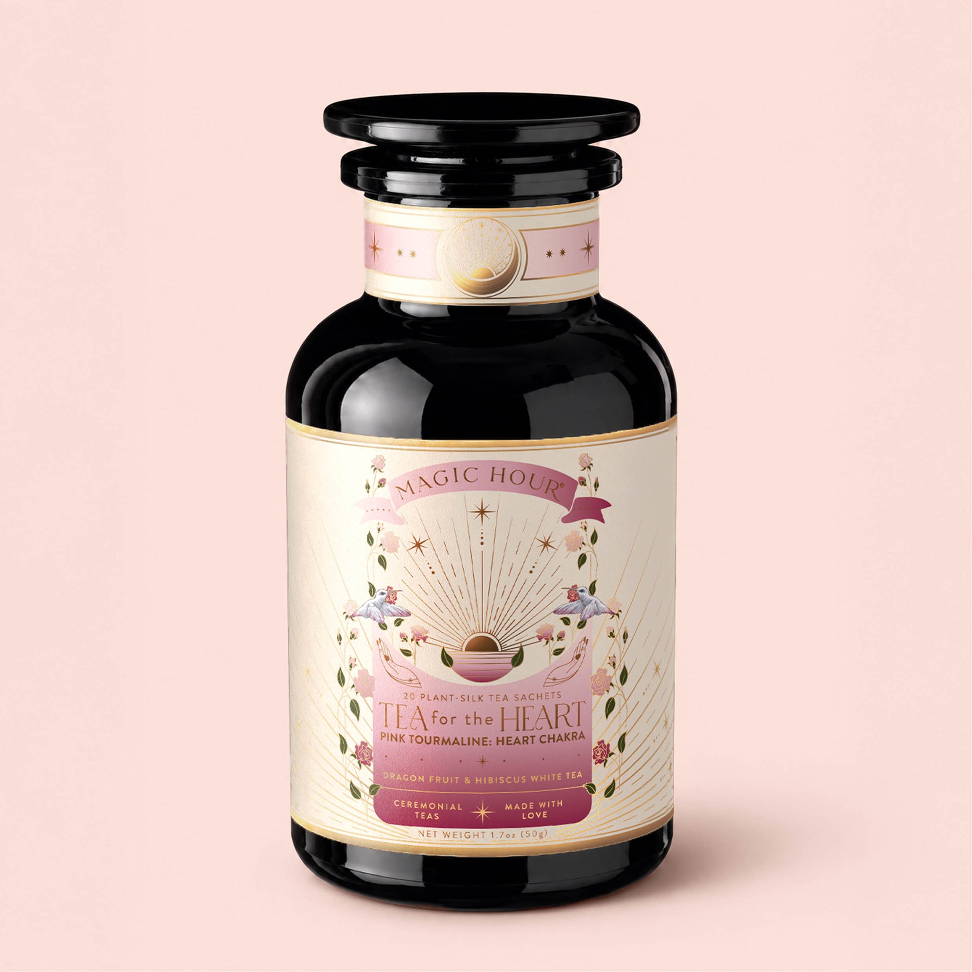 A black glass jar labeled "Tea for the Heart: Pink Tourmaline: Dragonfruit &amp; Hibiscus White Tea" by Magic Hour, adorned with pink, gold, and white designs featuring a sunburst, floral accents, and a heart. The label highlights that it’s a handcrafted heart chakra tea blended with delicate pink white tea and nourishing adaptogenic herbs.