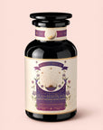 A dark glass jar labeled "Tea for Harmony: Queen of Wellness: Women's Cycle Herbal Blend" with purple, gold, and white design elements—including sun rays and flowers. The jar features a wide black lid and has a decorative, mystical appearance—perfect for women's herbal tea blends such as raspberry leaf or soothing organic chaste berry infusions.