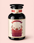 A glass jar labeled "Tea for Manifesting: Ruby Moon: Hibiscus, Elderberry & Passionfruit Herbal Tea" features enchanting illustrations of plants, glowing rays, and a crescent moon set against a pink background. The design exudes a sense of harmony and balance, capturing the essence of manifestation from root to crown chakra.