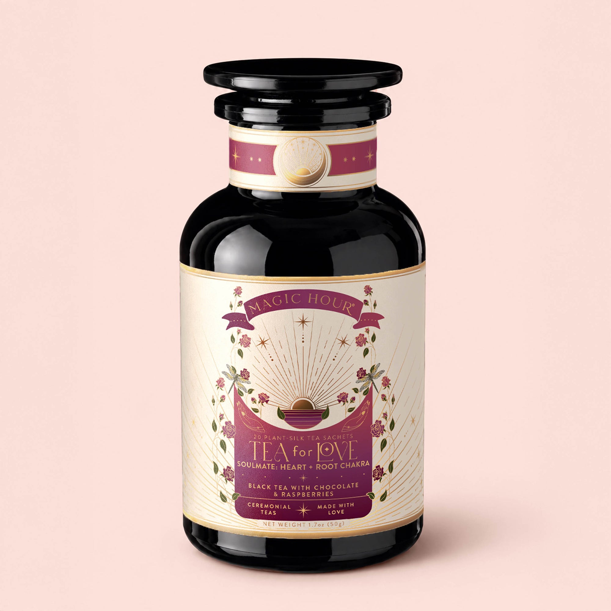 A dark glass jar with a gold and pink label reading "Tea for Love: Soulmate" from Magic Hour, adorned with stars, leaves, and sunburst designs, sits on a light pink background. This enchanting Black Tea with Chocolate &amp; Raspberries blends hand-selected Fair Trade leaves and stunning artwork to celebrate every pour.