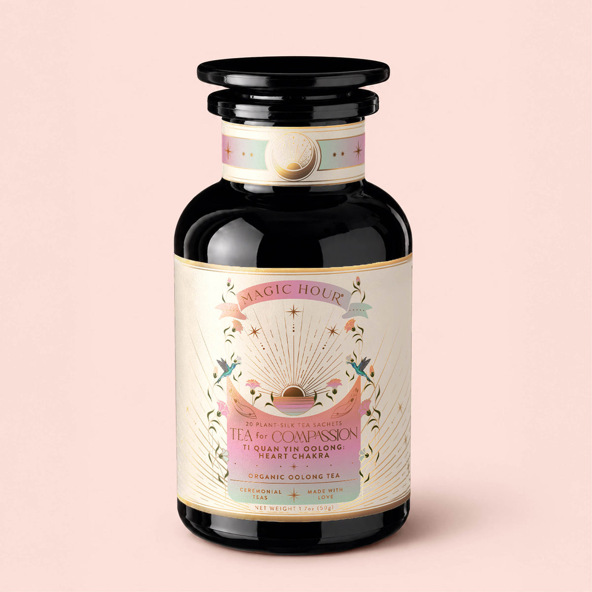 A glass jar of Magic Hour’s "Tea for Compassion: Ti Quan Yin: Organic Oolong Tea," adorned with pink, gold, and white celestial-themed artwork, containing organic oolong tea sachets.
