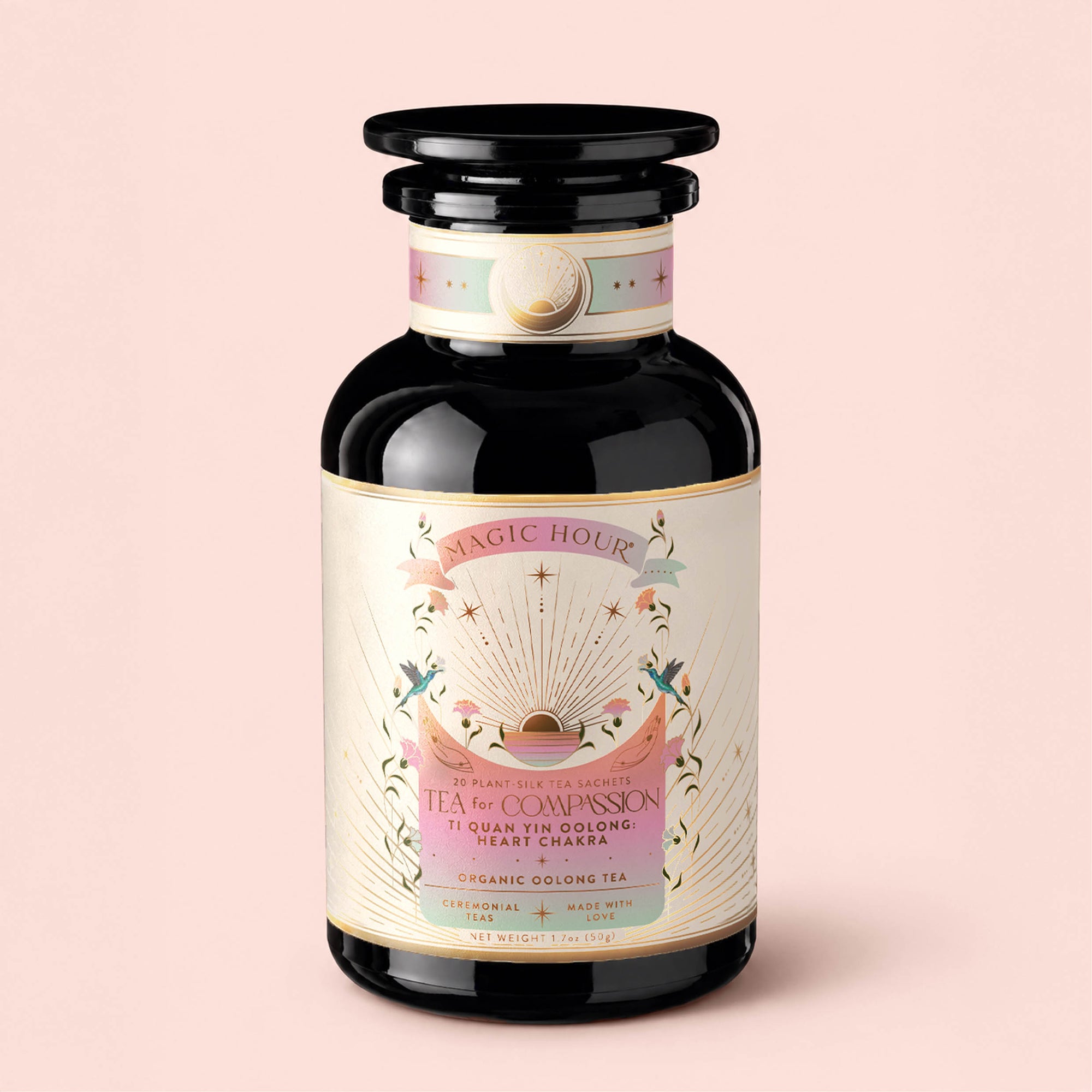 A glass jar of Magic Hour’s "Tea for Compassion: Ti Quan Yin: Organic Oolong Tea," adorned with pink, gold, and white celestial-themed artwork, containing organic oolong tea sachets.