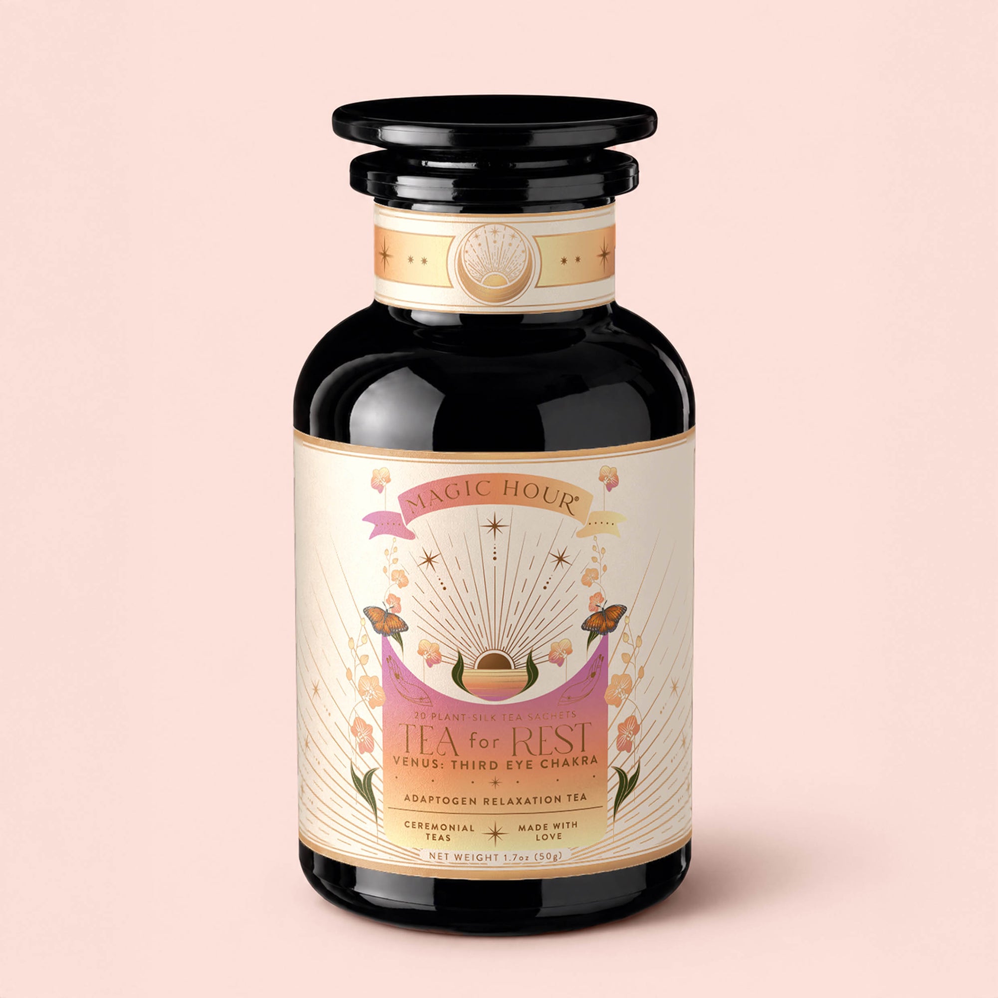 A dark glass jar labeled “Tea for Rest: Venus: Mango Blossom &amp; Nervines Herbal Tea,” adorned with floral designs, gold accents, and a decorative lid, is set against a pale pink background. Infused with an organic mango blossom and nervines herbal blend, this tea is thoughtfully crafted to support relaxation and promote natural collagen production.