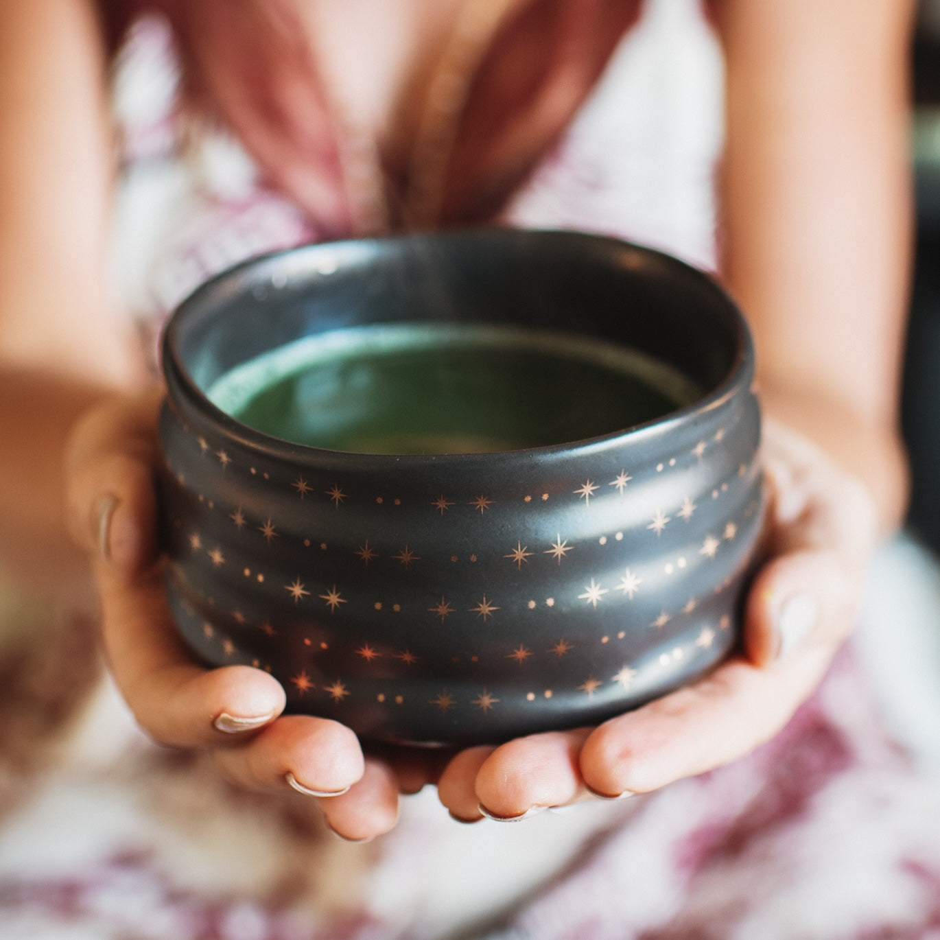 A person in a patterned dress holds the Tea Ceremony Starter Set’s elegant black bowl with star designs, filled with steaming green liquid. The emphasis is on the set and hands, while the background stays softly blurred.