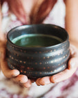 A person in a patterned dress holds the Tea Ceremony Starter Set’s elegant black bowl with star designs, filled with steaming green liquid. The emphasis is on the set and hands, while the background stays softly blurred.