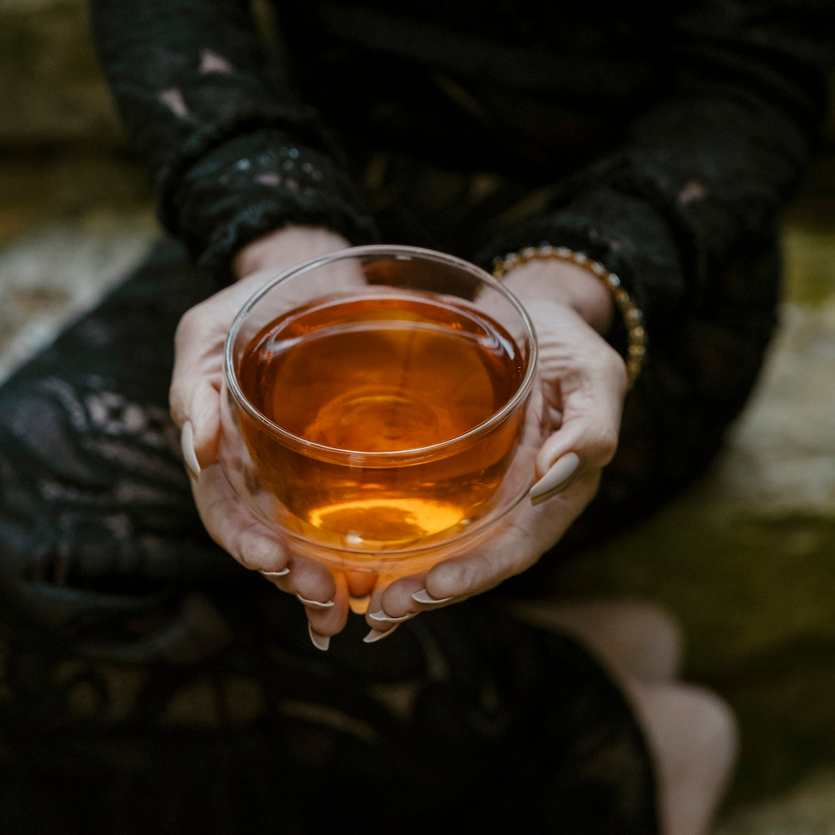 Wearing black lace, a person holds a clear glass of amber tea—possibly from our Small Batch Halloween Bundle—resting it on their lap.
