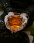 Wearing black lace, a person holds a clear glass of amber tea—possibly from our Small Batch Halloween Bundle—resting it on their lap.