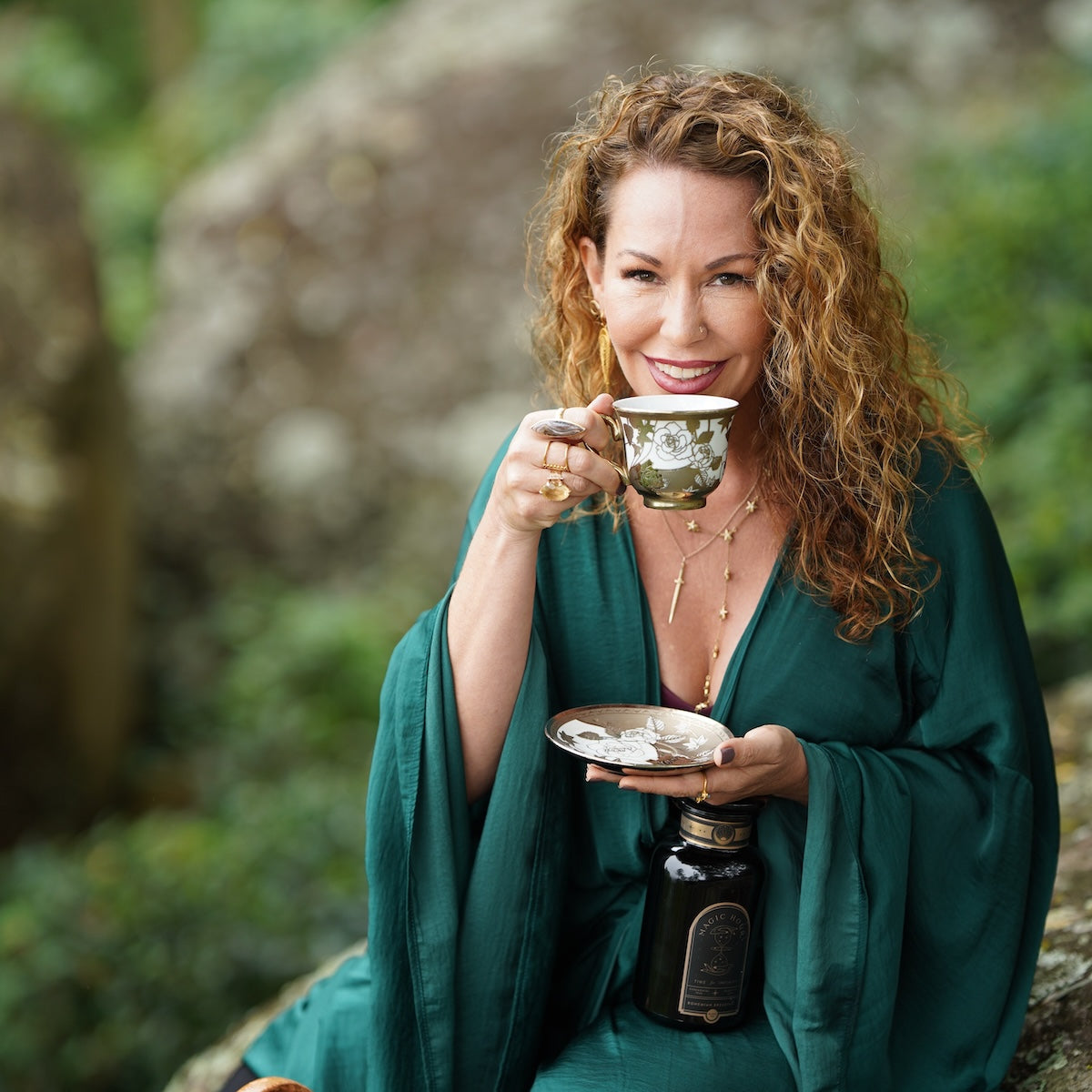 A woman with curly hair, wearing a deep green robe, holds a decorative teacup and saucer while smiling at the camera. As a Master Tea Blender representing Magic Hour, she is seated outdoors in a natural setting with blurred greenery in the background, also holding a black bottle in her lap. She embodies the essence of tea expertise taught in the Tea School 2024-2025 Full Course Bundle: Unlock the Secrets of Tea Mastery.