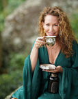 A woman with curly hair, wearing a deep green robe, holds a decorative teacup and saucer while smiling at the camera. As a Master Tea Blender representing Magic Hour, she is seated outdoors in a natural setting with blurred greenery in the background, also holding a black bottle in her lap. She embodies the essence of tea expertise taught in the Tea School 2024-2025 Full Course Bundle: Unlock the Secrets of Tea Mastery.