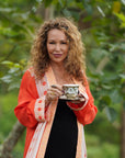 A woman with curly hair, draped in a vibrant orange and white floral-patterned robe, stands outdoors in a lush, green garden. She holds a teacup and saucer, smiling warmly at the camera, fully embodying the expertise of a Master Tea Blender from Magic Hour’s esteemed Tea School 2024-2025 Full Course Bundle: Unlock the Secrets of Tea Mastery.