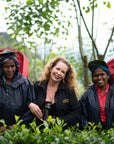 A person with curly hair in a black jacket stands in a tea plantation, smiling and holding "Tea School Course 