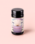 A canister of Tea for Healing: Aquamarine Dream—Blueberries, Pineapple & Nervines Herbal Tea—stands on a pastel pink background. The label showcases botanical illustrations, colorful details, and highlights the presence of butterfly pea flowers alongside notes of pineapple. This adaptogenic blend is carefully crafted to support the nervous system and promote overall wellness.