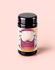 A black tin labeled “Tea for Celebration: Coconut Chai: Rich Spiced Black Tea Blend” fair trade spiced black tea sits on a pale pink background. The label features floral designs and an open book with rays emanating from it.