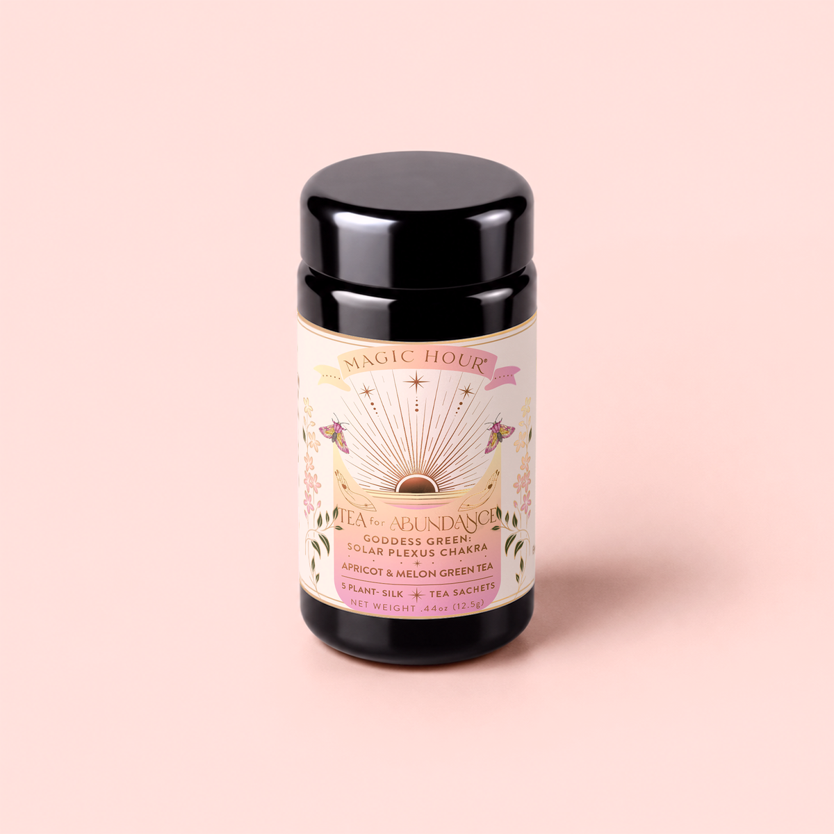 A black tea canister labeled “Magic Hour” rests on a pink surface. The label showcases colorful floral designs, a rainbow, and the words “Tea for Abundance: Goddess Green: Apricot & Lychee Green Tea.” Infused with green tea and organic lemongrass to help support metabolism, this blend delivers vibrant flavor and uplifting energy.