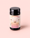 A black tea canister labeled “Magic Hour” rests on a pink surface. The label showcases colorful floral designs, a rainbow, and the words “Tea for Abundance: Goddess Green: Apricot & Lychee Green Tea.” Infused with green tea and organic lemongrass to help support metabolism, this blend delivers vibrant flavor and uplifting energy.