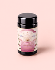 A canister of “Tea for the Heart: Pink Tourmaline – Dragonfruit & Hibiscus White Tea” rests on a pale pink background. The label, adorned with floral motifs, two doves, and radiant sun rays, highlights the blend’s hibiscus fruit and adaptogenic herbs. Subtle design elements hint at the delicate allure of dragonfruit and pink white tea.