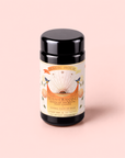 A black canister labeled “Tea for Cleansing: Queen of the South: Herbal Cafe de Olla” rests on a pale pink background. The label displays floral designs, two hummingbirds, and celestial motifs, with text highlighting its antioxidant-rich and caffeine-free herbal cafe de olla tea sachets.