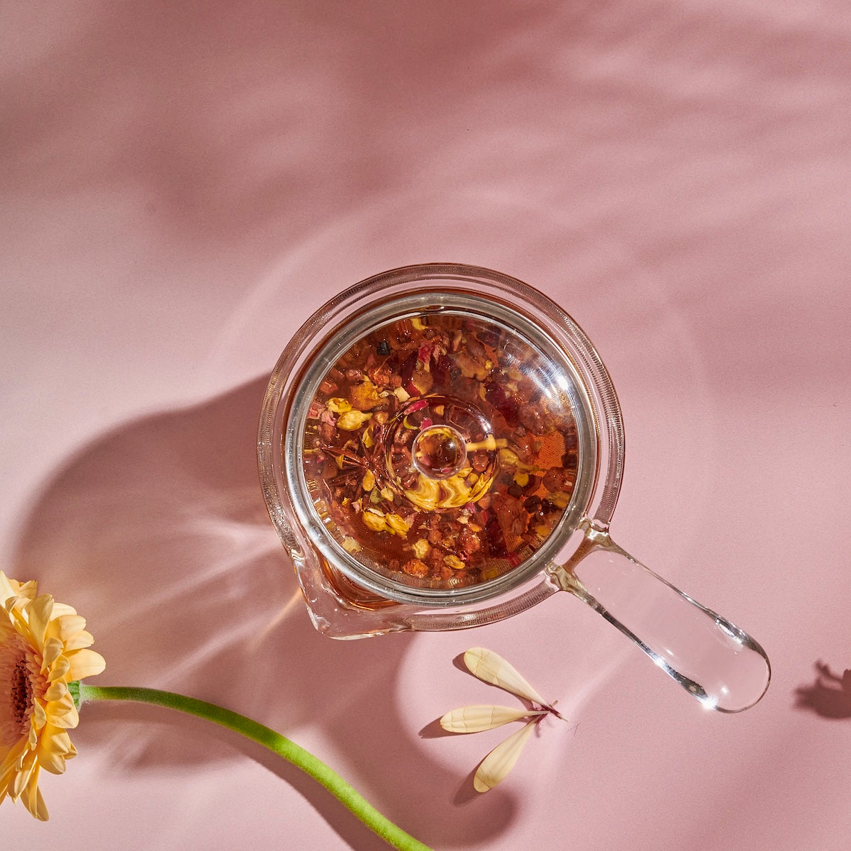 A glass Tea in Hand Teapot from The Best of Magic Hour Sampler Set filled with herbal tea, viewed from above next to a yellow flower on a pink surface.